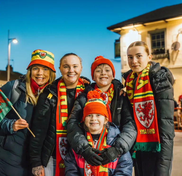 A family of Welsh football fans wearing Wales kit and smiling at the camera