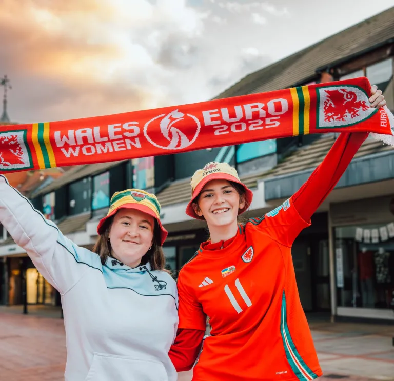 Two people holding a 'WALES WOMEN EURO 2025' scarf in a shopping area, with shops and a cloudy sky behind them.