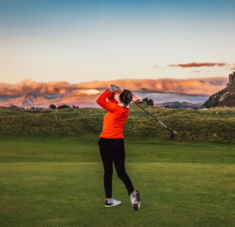 Golfer mid-swing on a lush course, backed by scenic hills and a historic castle atop a hill beneath a vibrant, cloud-filled sky.