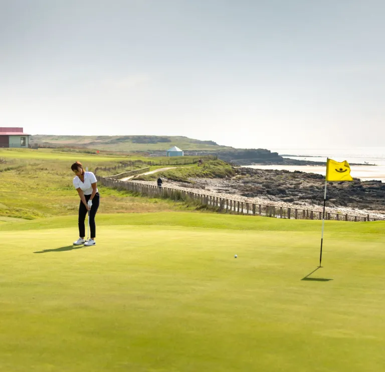 A golfer stood on a green putting towards the hole with a flag in it. The club building can be seen in thew distance behind.