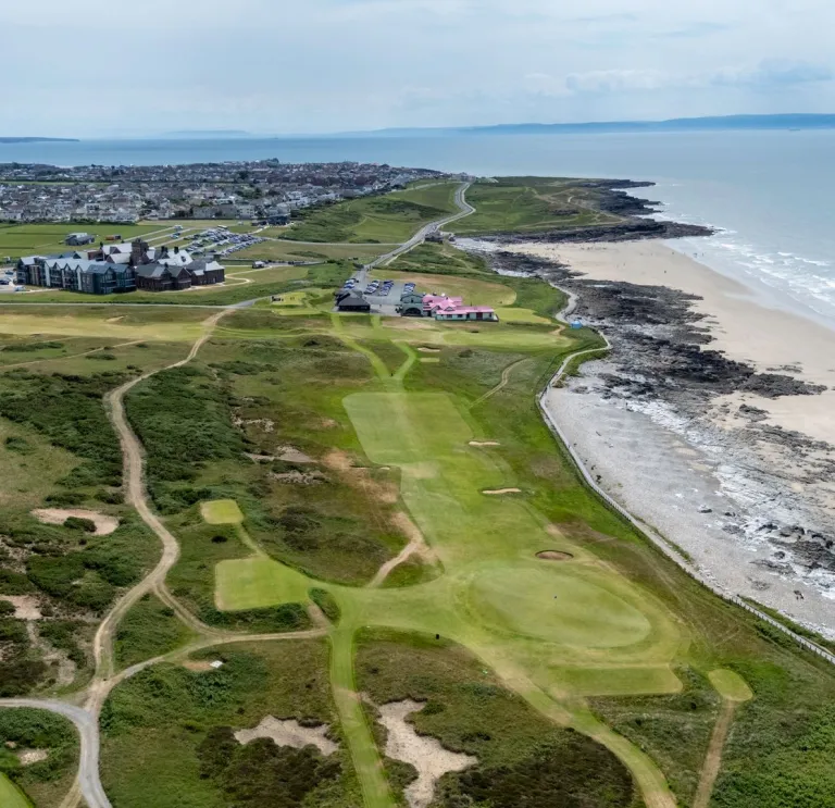 Aerial view of a coastal golf course with green fairways, a sandy beach, and rocky shoreline on the right, and a town in the background.