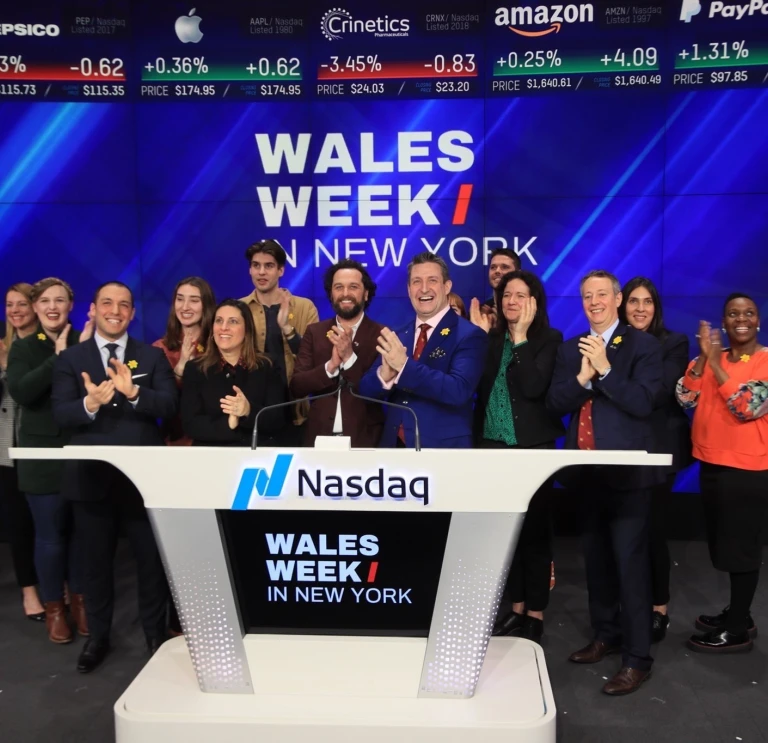 A group of people stand on the Nasdaq stage clapping and smiling during a Wales Week in New York event, with stock tickers displayed above them.