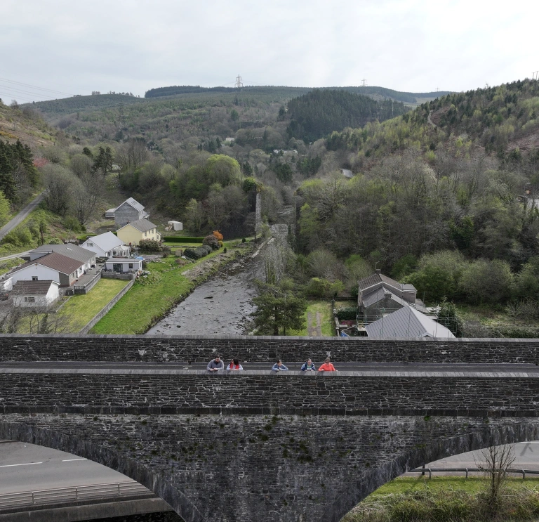 A wide view of a stone bridge with several people standing on it, overlooking a valley with houses, trees and a river running below.