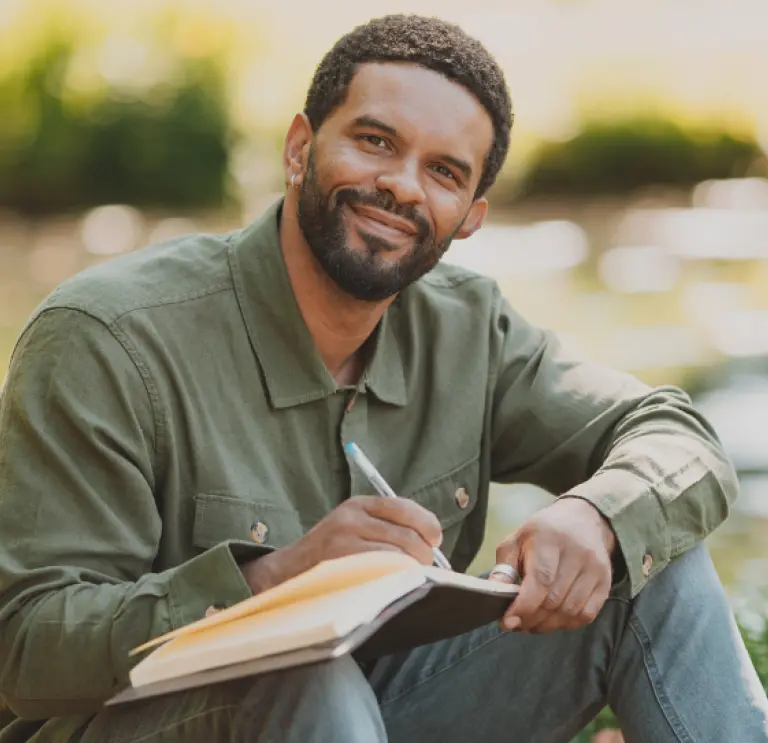 Alex Wharton sitting by a stream smiling with a book on his lap