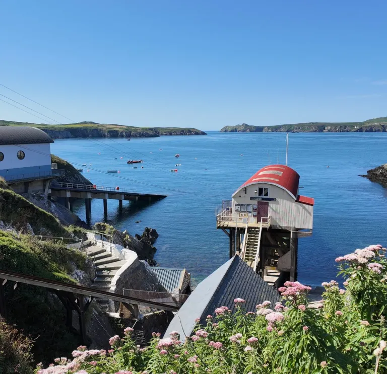 Un petit port côtier aux eaux bleues et calmes, avec un canot de sauvetage à toit rouge amarré près d’une rampe de mise à l’eau, des bâtiments blancs le long du rivage, et des falaises verdoyantes encadrant la scène sous un ciel dégagé.
