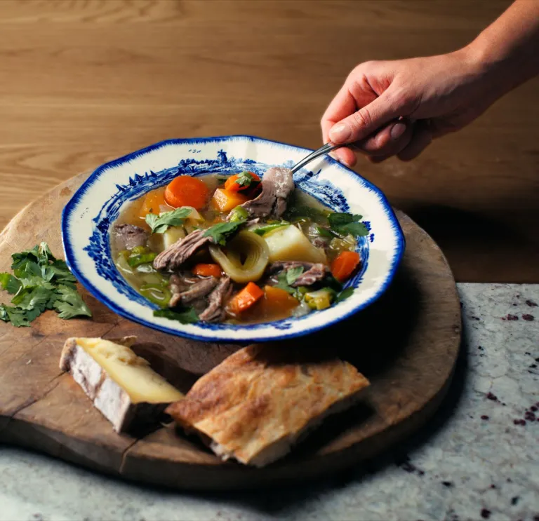 Ein Bild einer schön präsentierten Schüssel walisischen Cawl auf einem Holzbrett, daneben Käse und Brot.