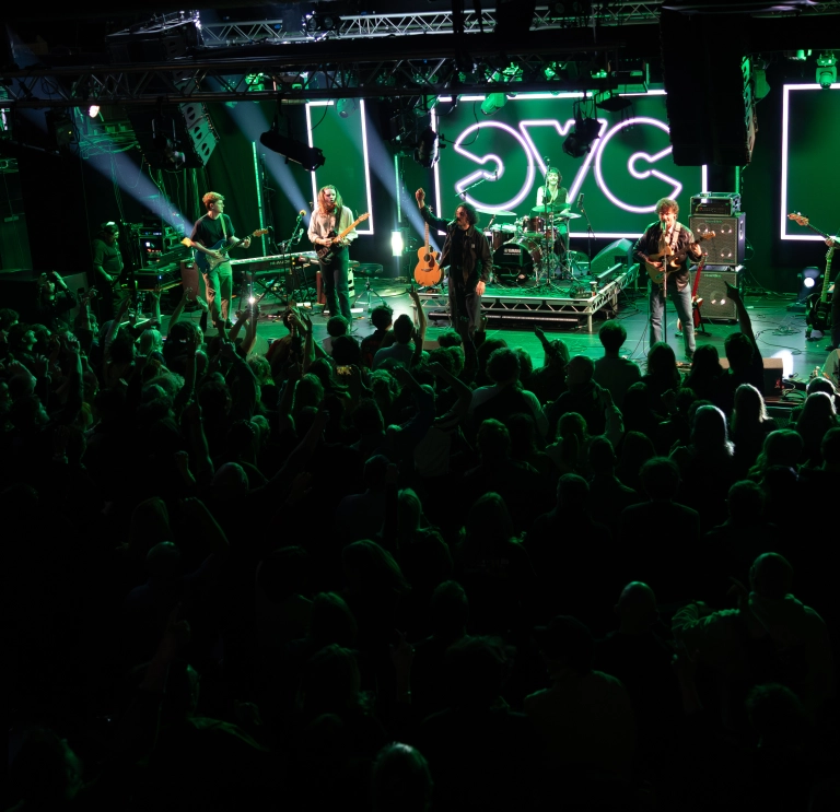 A band performing on stage in front of a large crowd. Bright green lights illuminate the scene.