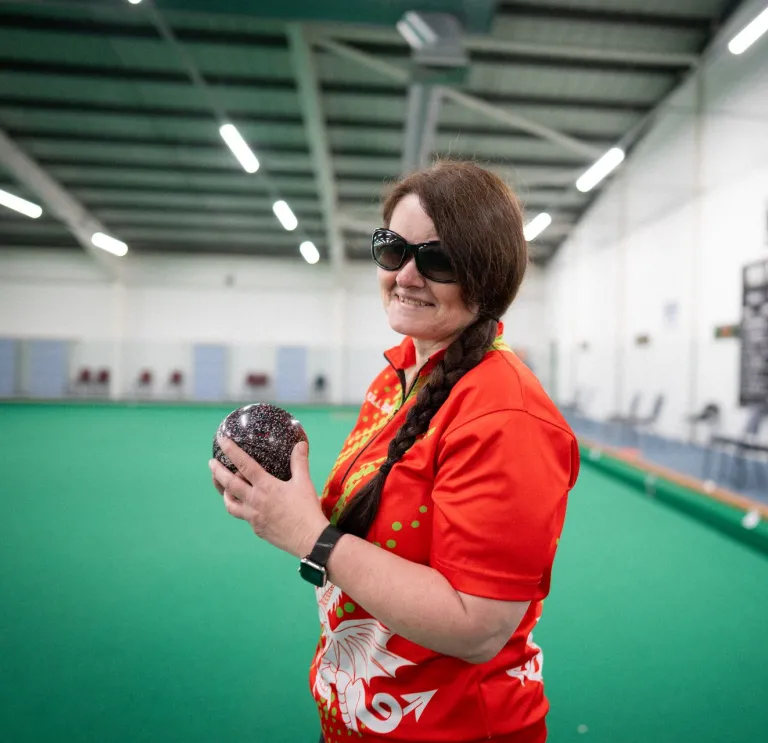 Julie Thomas holding a bowls ball on an indoor bowling green for Team Wales.