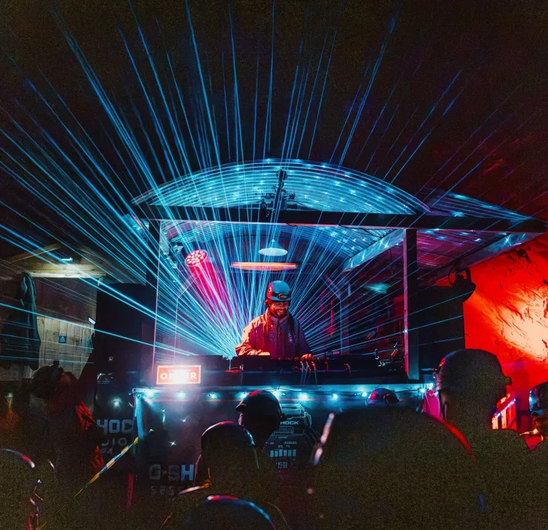 DJ Roni Size performing beneath blue laser lights at an underground rave in a red-lit cavern. 