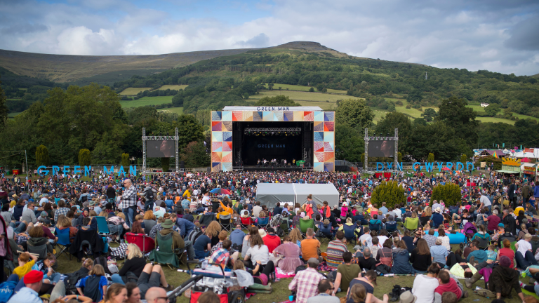 A crowd of people sat in a field looking towards an outdoor festival stage with a hill in the background