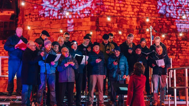 A male voice choir singing in front of a castle that is lit up in red 