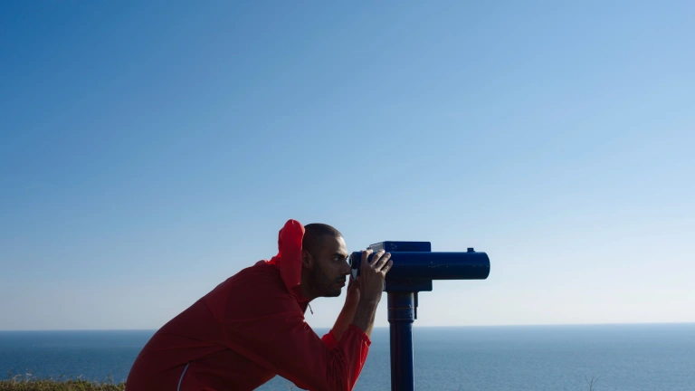 Side on view of a man in a red coat bending down to look through a telescope pointed out of right of frame.