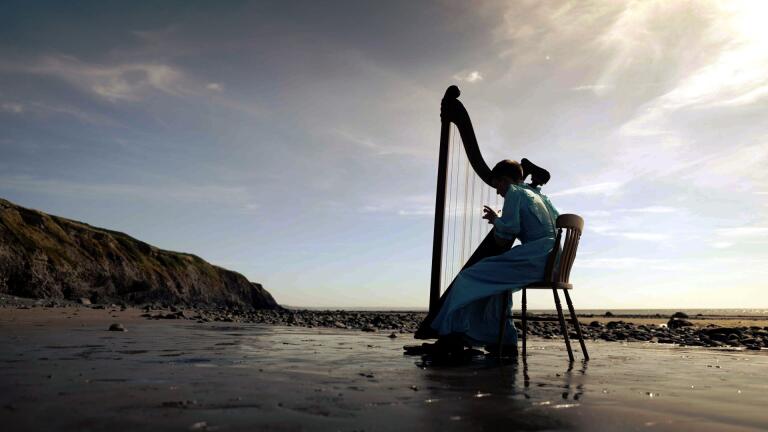 A young harp player wearing a long dress, sitting and playing a harp on a beautiful deserted beach