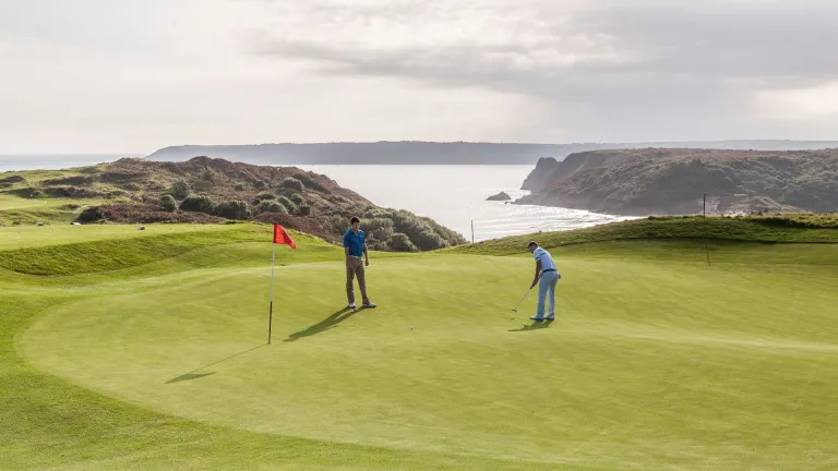 Two people playing golf on the green with views of the sea and mountains in the background.