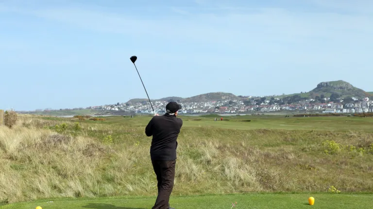 Golfer in black mid-swing on a lush green course with hills and a town in the background under a clear blue sky.