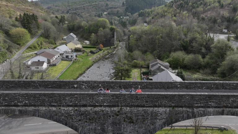 A wide view of a stone bridge with several people standing on it, overlooking a valley with houses, trees and a river running below.