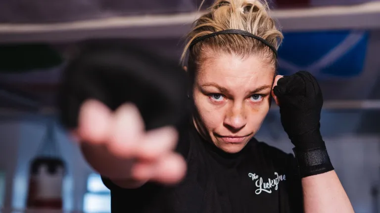 Lauren Price MBE in a boxing gym, wearing black training gear with her gloves raised, extending one fist towards the camera during a training session. 