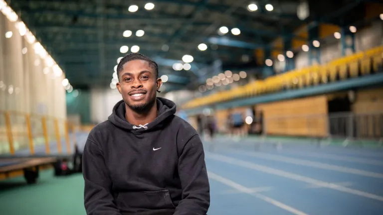 Jeremiah Azu sitting on an indoor athletics track, smiling at the camera.