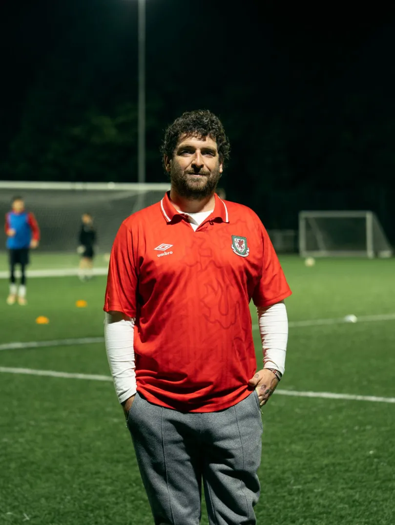 Club founder Delwyn Derrick standing on a football pitch while a game takes places behind him