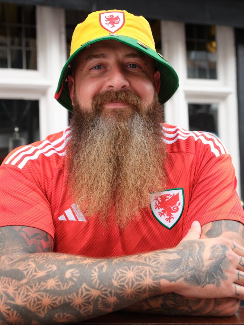 A man with a long beard and tattooed arms smiling at the camera, while wearing a Welsh bucket hat and Wales football strip