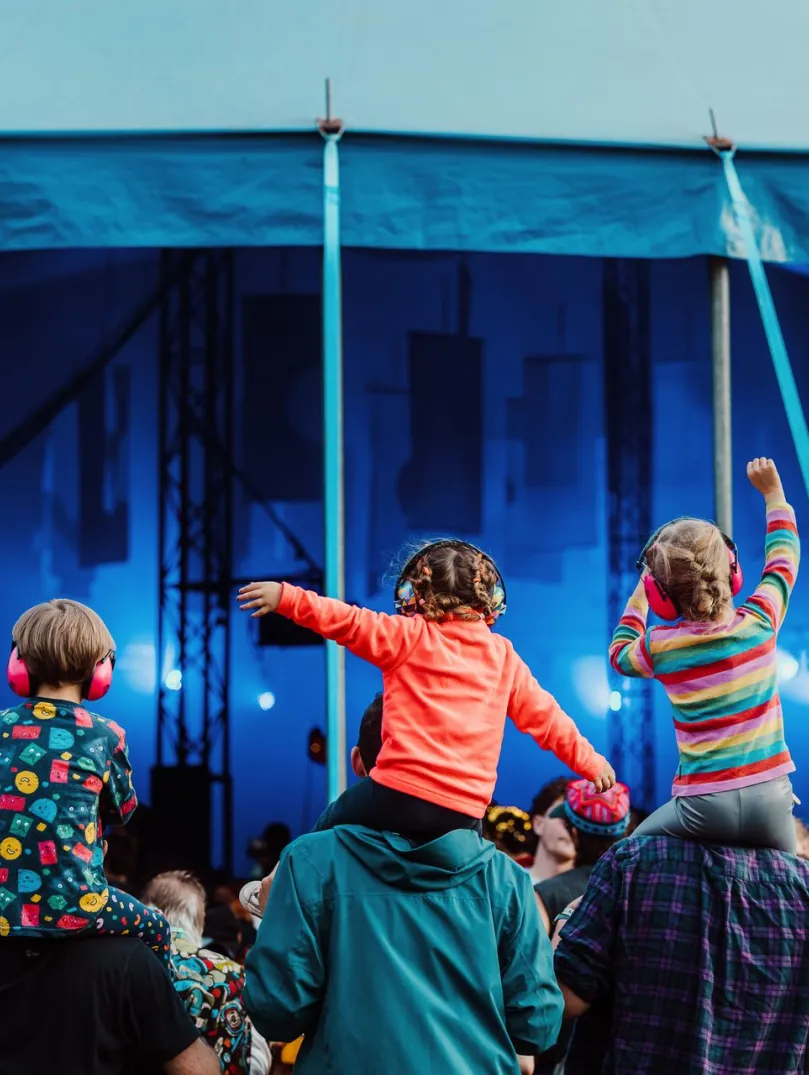 Three children sat on the shoulders of their parents listening to a band playing at a festival.