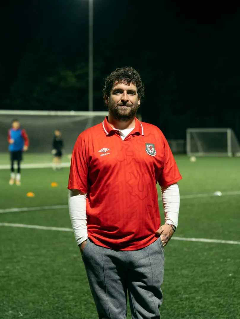 Le fondateur du club, Delwyn Derrick, debout sur un terrain de football, tandis qu'un match se déroule en arrière-plan.