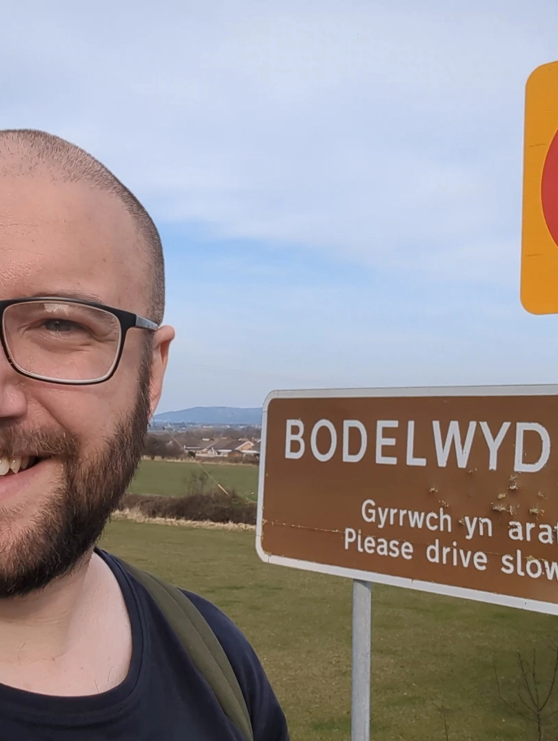 Joe taking a selfie beside a “Bodelwyddan” village sign in a grassy landscape.