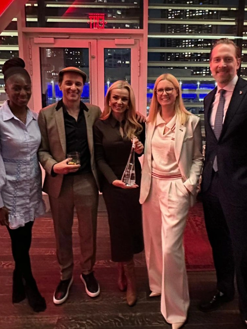 A group of five people pose together at an indoor event with city lights visible through the windows.