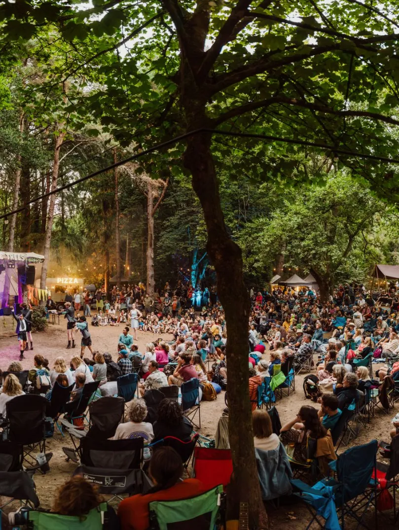A large outdoor audience seated on folding chairs in a woodland clearing, watching a live performance on a small stage lit with purple lights, with trees overhead, food stalls in the background, and a relaxed festival atmosphere.