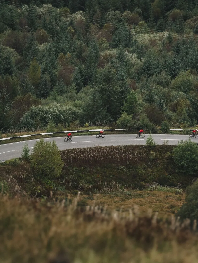 Four cyclists in red jerseys cycling round a tight bend in a mountainous road, surrounded by a mixture of beautiful trees