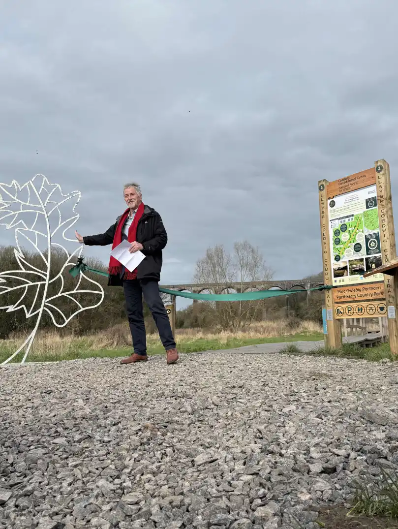 A man speaks beside a leaf shaped metal sculpture and a green ribbon across a gravel path at a country park entrance, with others watching and a viaduct in the background.