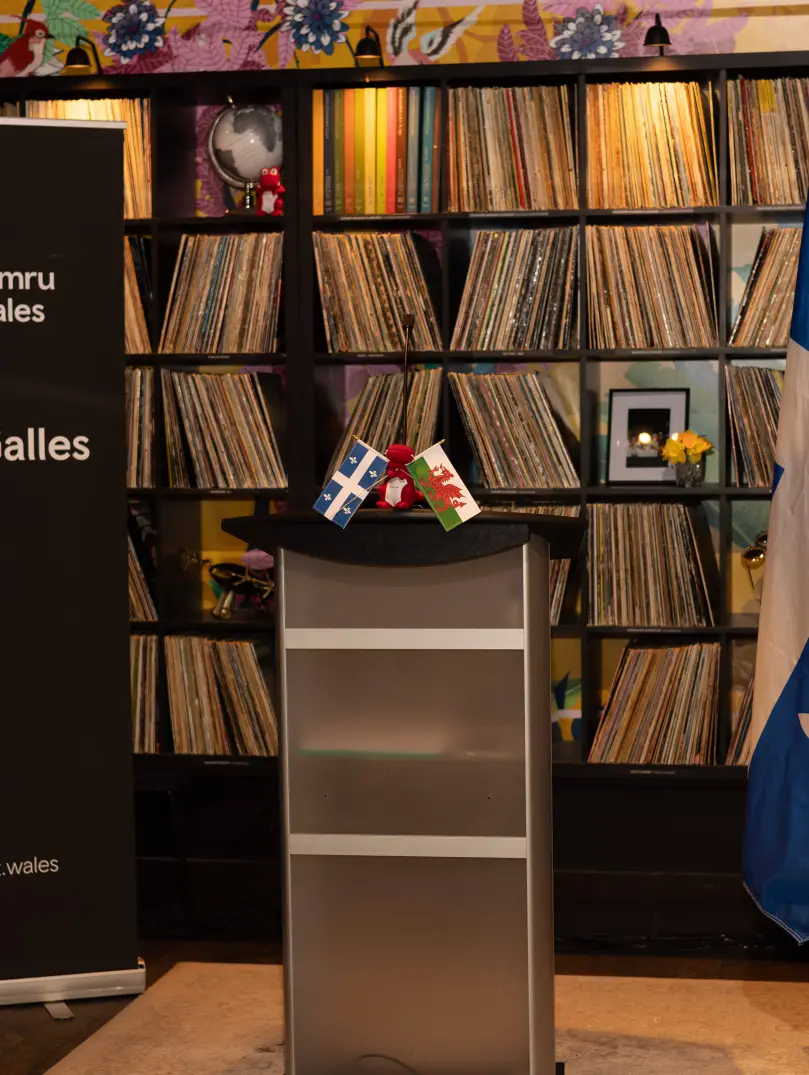 Podium with small Welsh and Québec flags, beside a Cymru Wales “Pays de Galles” banner, with full sized Québec and Wales flags and shelves of vinyl records behind.