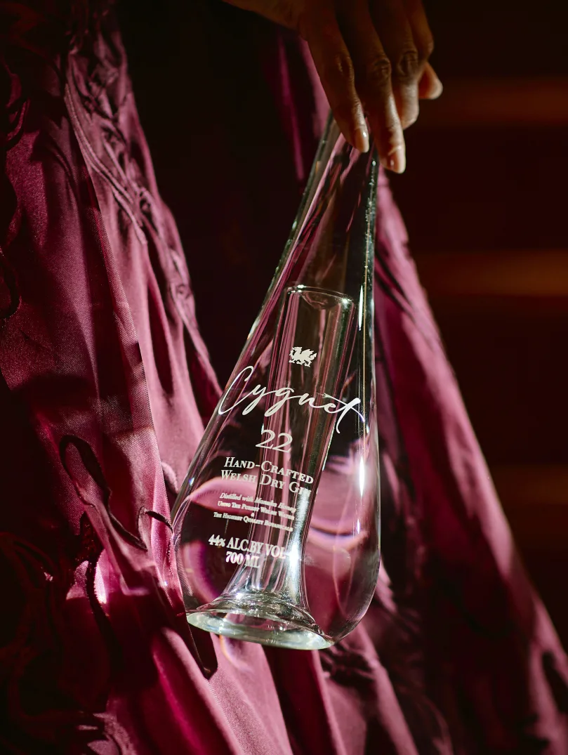 Hand holding an engraved glass gin bottle against a red satin dress in warm light.