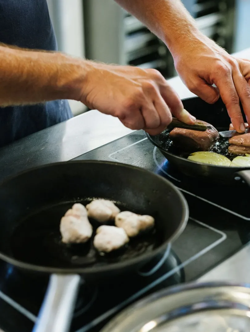 A person cooking food in two frying pans on a hob, turning pieces of meat while onions cook in one pan.
