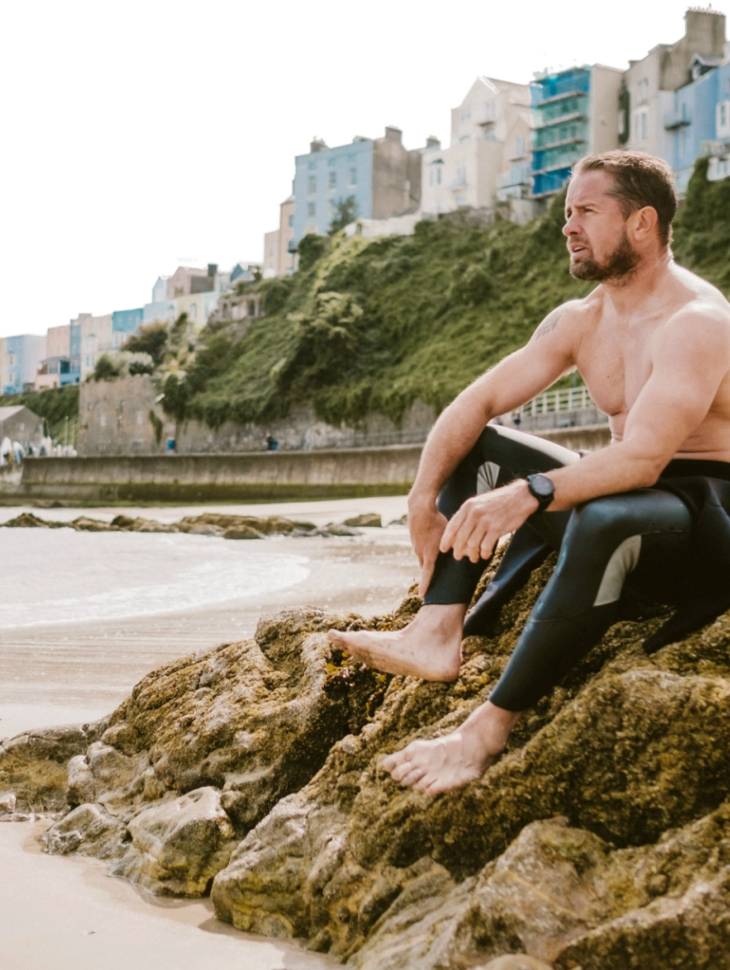 Shane Williams sitzt auf einem Felsen am North Beach in Tenby mit bunten Stadthäusern im Hintergrund.