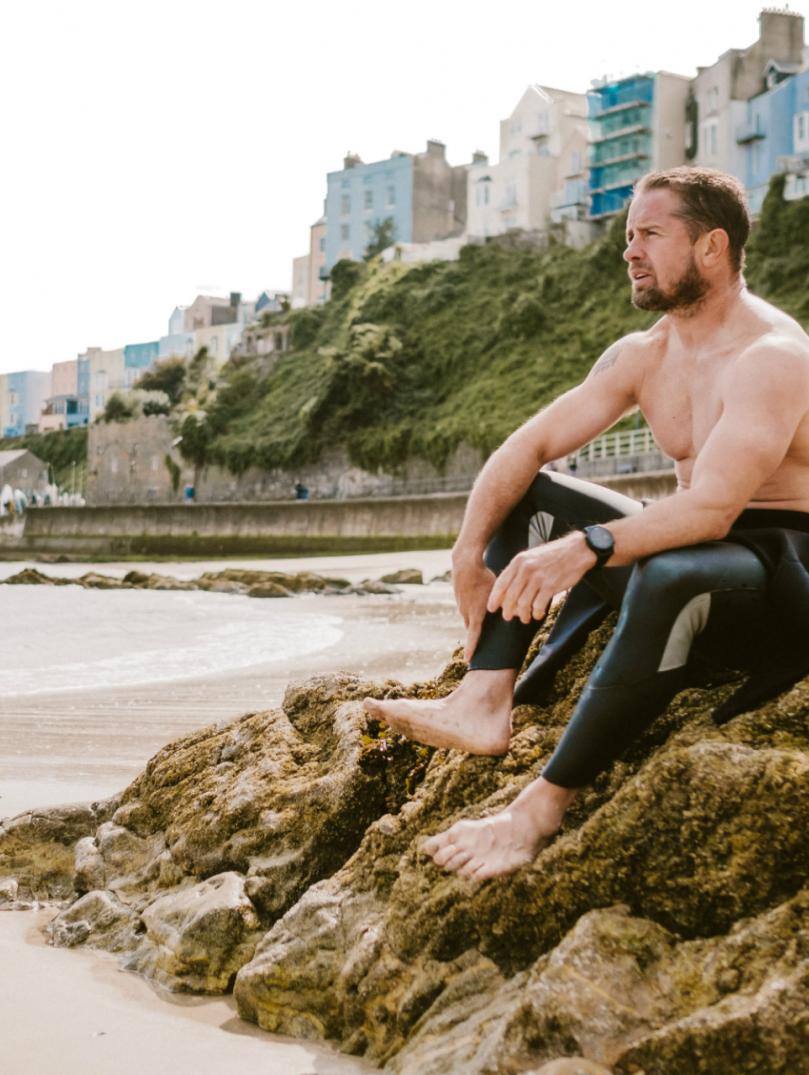 Shane Williams sitting on a rock on Tenby's North beach with multicoloured townhouses behind.