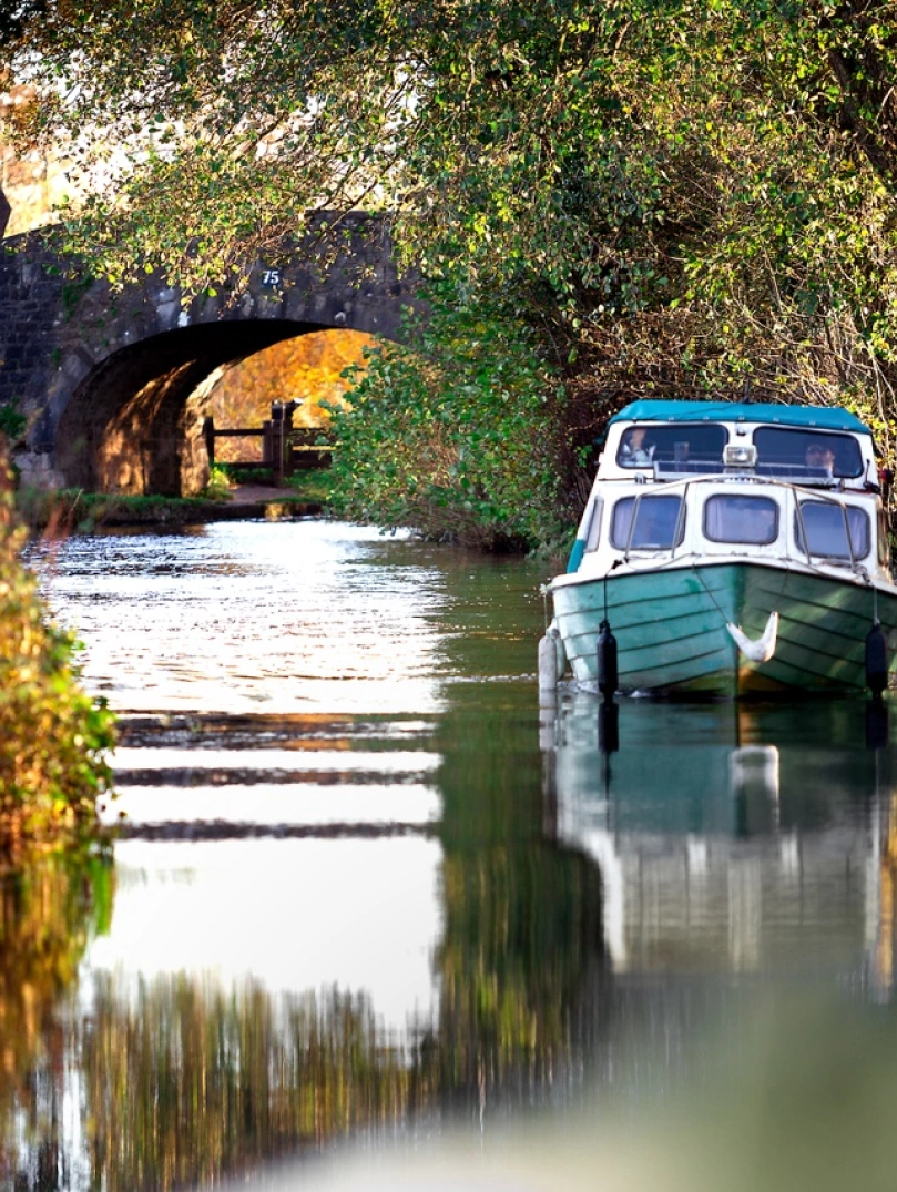 Hausboot auf einem Kanal umgeben von Bäumen, mit einer Brücke aus Stein im Hintergrund.