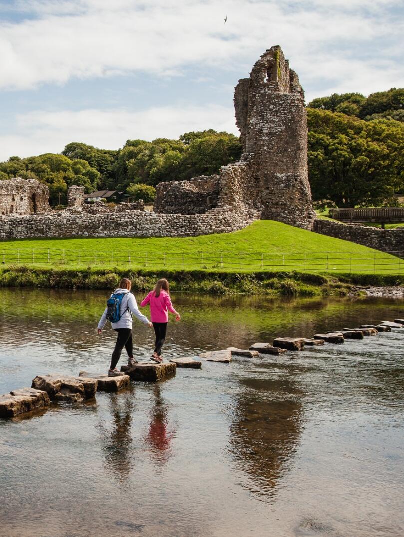 Caballos y caminantes frente a un castillo en ruinas rodeado de campos verdes y agua.