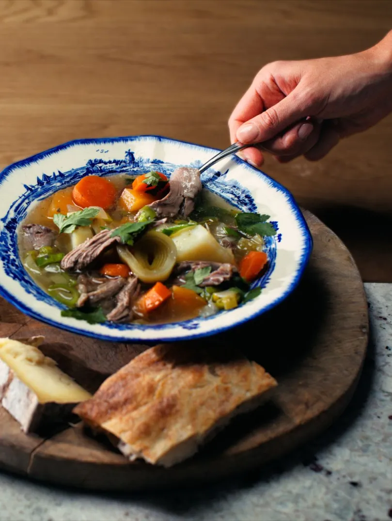 An image of a beautifully presented bowl of Welsh cawl on a wooden board with cheese and bread alongside