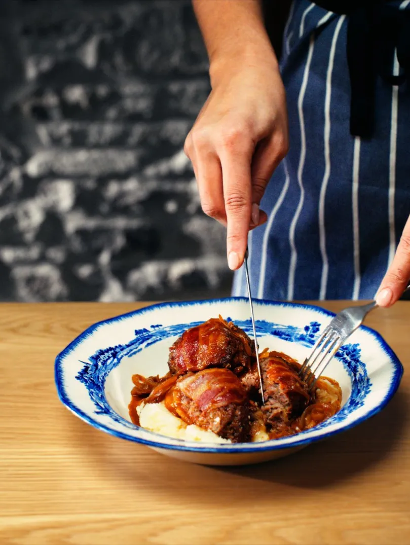 A beautifully presented bowl of faggots with mash, covered in gravy, being cut open for serving by a chef's hands