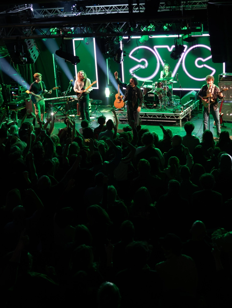 A band performing on stage in front of a large crowd. Bright green lights illuminate the scene.