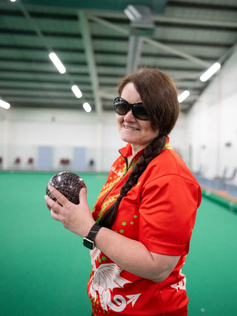 Julie Thomas holding a bowls ball on an indoor bowling green for Team Wales.