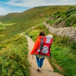 A parent with a baby in a hiking carrier walking down a beautiful coastal path with green to the right and the beach and blue sea to the left