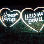 Projected images on a stone church wall - hearts with 'Other voices' and 'Lleisiau Eraill' text within.