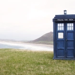A blue phone box sat on grass with the sea in background.