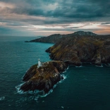 Dramatic coastal shot showing a lighthouse on exposed outlying rocky land, with blue seas and waves all around