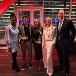 A group of five people pose together at an indoor event with city lights visible through the windows.