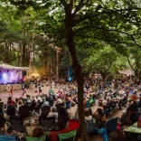 A large outdoor audience seated on folding chairs in a woodland clearing, watching a live performance on a small stage lit with purple lights, with trees overhead, food stalls in the background, and a relaxed festival atmosphere.