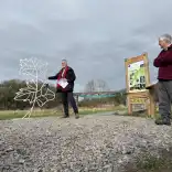 A man speaks beside a leaf shaped metal sculpture and a green ribbon across a gravel path at a country park entrance, with others watching and a viaduct in the background.