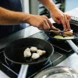 A person cooking food in two frying pans on a hob, turning pieces of meat while onions cook in one pan.
